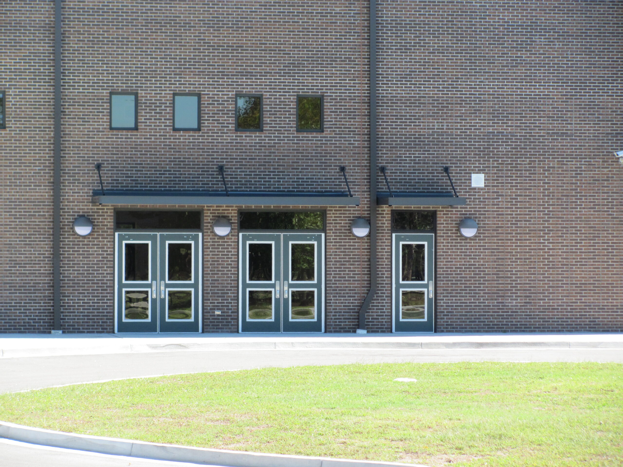 Three glass double doors and four small windows on a brick building, with grass and pavement in front.