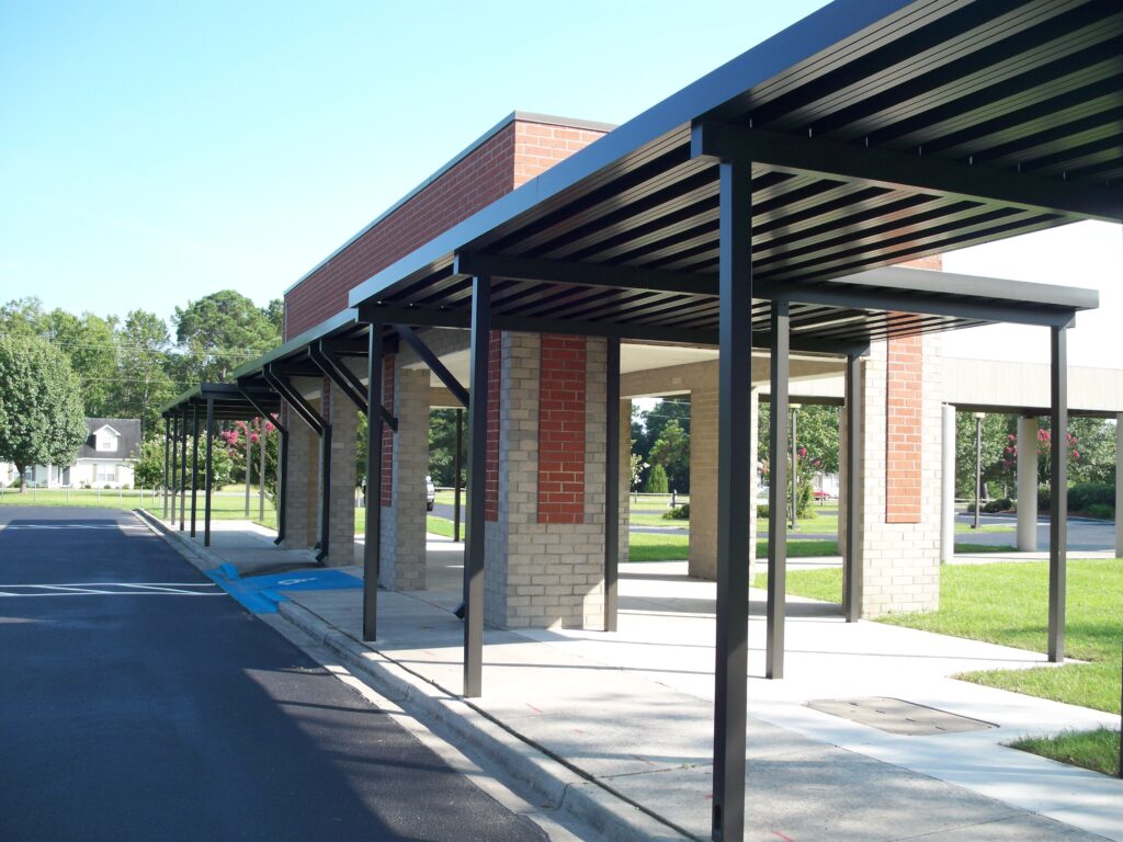 Covered walkway with metal roof and brick columns next to a paved road and grassy area on a sunny day.