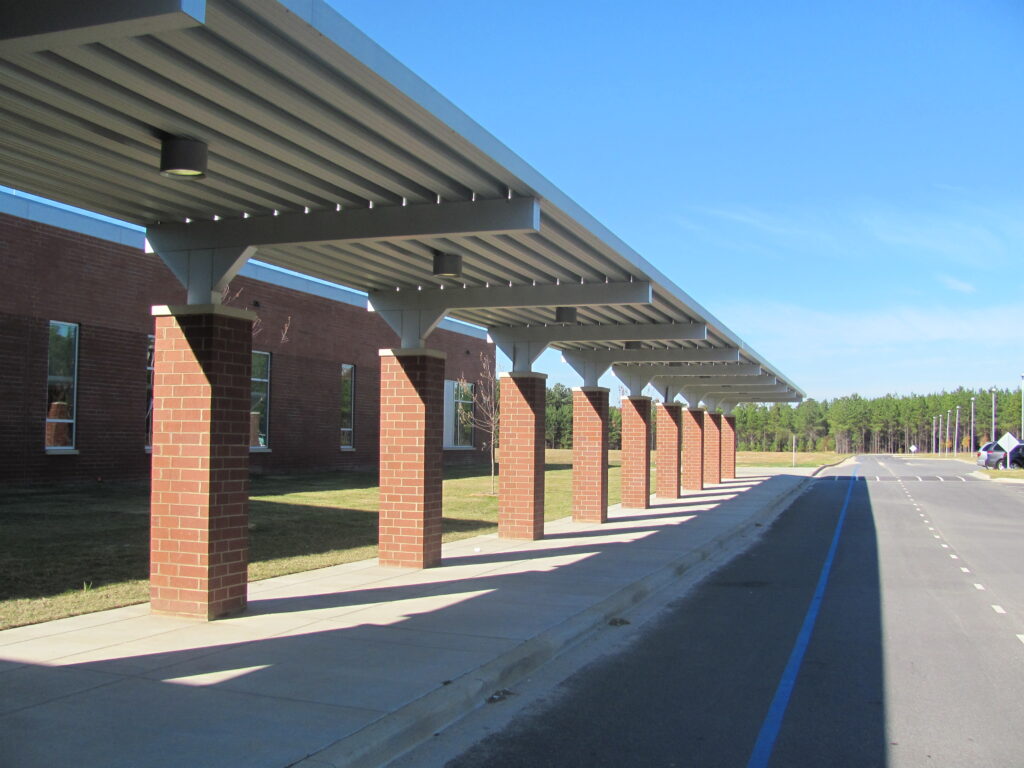 Covered walkway with brick pillars beside a brick building and an empty road under a clear blue sky.